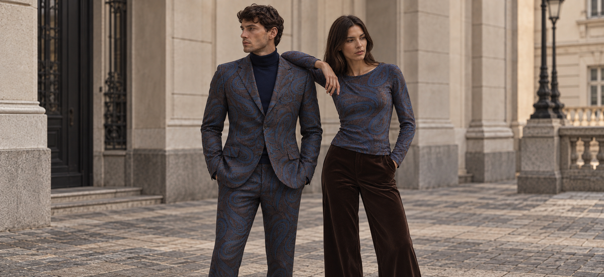 A man in Lords of Harlech Dragon paisley tailoring and a navy turtleneck beside a woman in brownie velvet trousers, in front of a classical stone facade