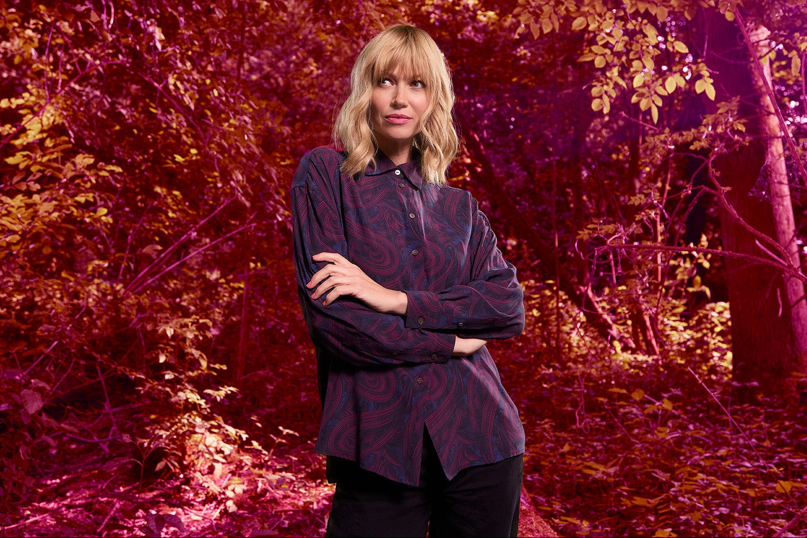 Woman in a plum paisley button down shirt standing in a forest with a red and purple color filter